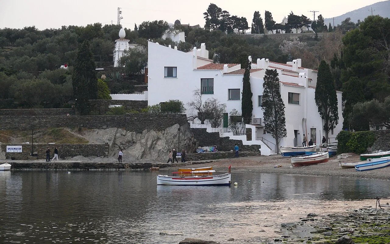 Dalís weiß getünchtes Haus in Portlligat vom Strand aus gesehen, Zypressen neben der Treppe und Fischerboote am Ufer