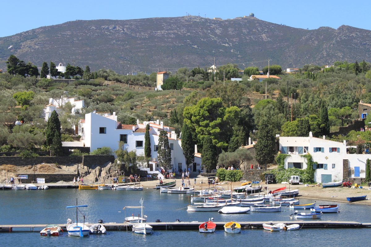 Salvador Dalís weiß getünchtes Haus in Portlligat mit weißen Eiern auf dem Dachfirst, über einer Bucht mit vertäuten Fischerbooten und den Hügeln des Cap de Creus